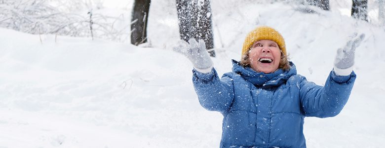 Winter scene of a beautiful smiling senior woman in a blue coat and white gloves with her hands raised toward falling snow.